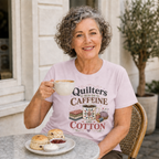 A smiling woman with grey hair sits at an outdoor cafe table holding a cup of coffee. She is wearing a pale mauve t-shirt with 'Quilters Run On Caffeine and Cotton' above an illustration of a coffee mug, fabric stack, thread spools and a pincushion. Scones with jam and cream are on the table in front of her.