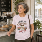 A smiling woman with curly gray hair stands in a coffee shop wearing a light mauve graphic T-shirt that reads 'Quilters Run On Caffeine and Cotton' with fabric stack, coffee mug, pincushion and thread spools. The background shows a cafe counter with pastries, glass jars, and a coffee machine.