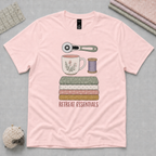 A light pink short-sleeve t-shirt laid flat on a gray cutting mat, featuring a central graphic of sewing and crafting essentials including a rotary cutter, mug, spool, folded fabrics, and the text 'RETREAT ESSENTIALS'.