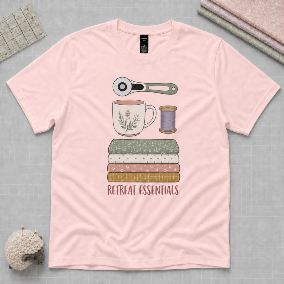 A light pink short-sleeve t-shirt laid flat on a gray cutting mat, featuring a central graphic of sewing and crafting essentials including a rotary cutter, mug, spool, folded fabrics, and the text 'RETREAT ESSENTIALS'.