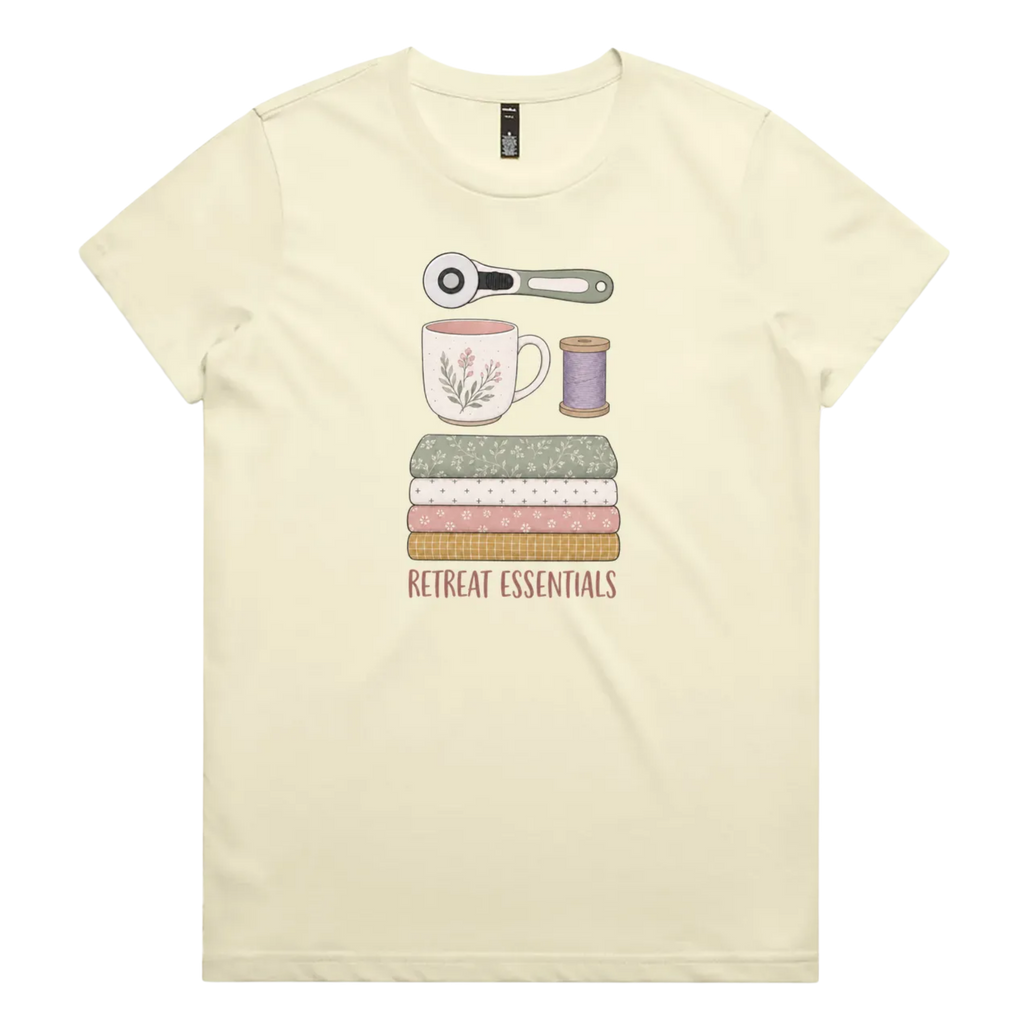 A light butter yellow short-sleeve crewneck t-shirt featuring a graphic of sewing and crafting essentials (a rotary cutter, mug, fabric stack, spool of thread) with the text 'RETREAT ESSENTIALS' printed below.