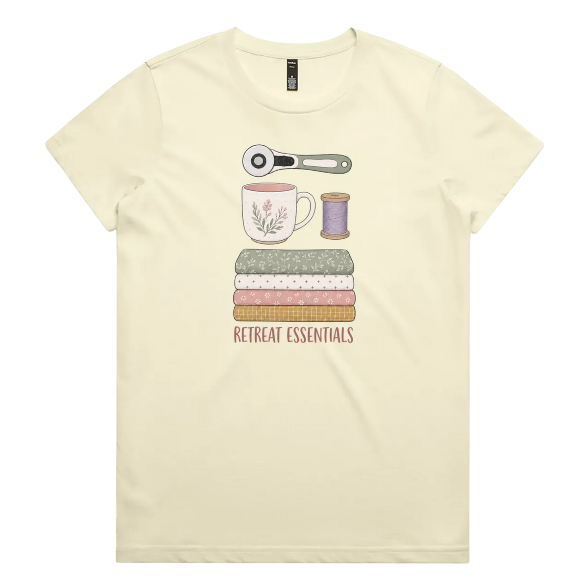 A light butter yellow short-sleeve crewneck t-shirt featuring a graphic of sewing and crafting essentials (a rotary cutter, mug, fabric stack, spool of thread) with the text 'RETREAT ESSENTIALS' printed below.