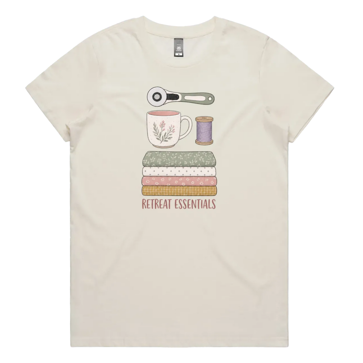 An off-white short-sleeve crewneck t-shirt featuring a graphic of sewing and crafting essentials (a rotary cutter, mug, fabric stack, spool of thread) with the text 'RETREAT ESSENTIALS' printed below.