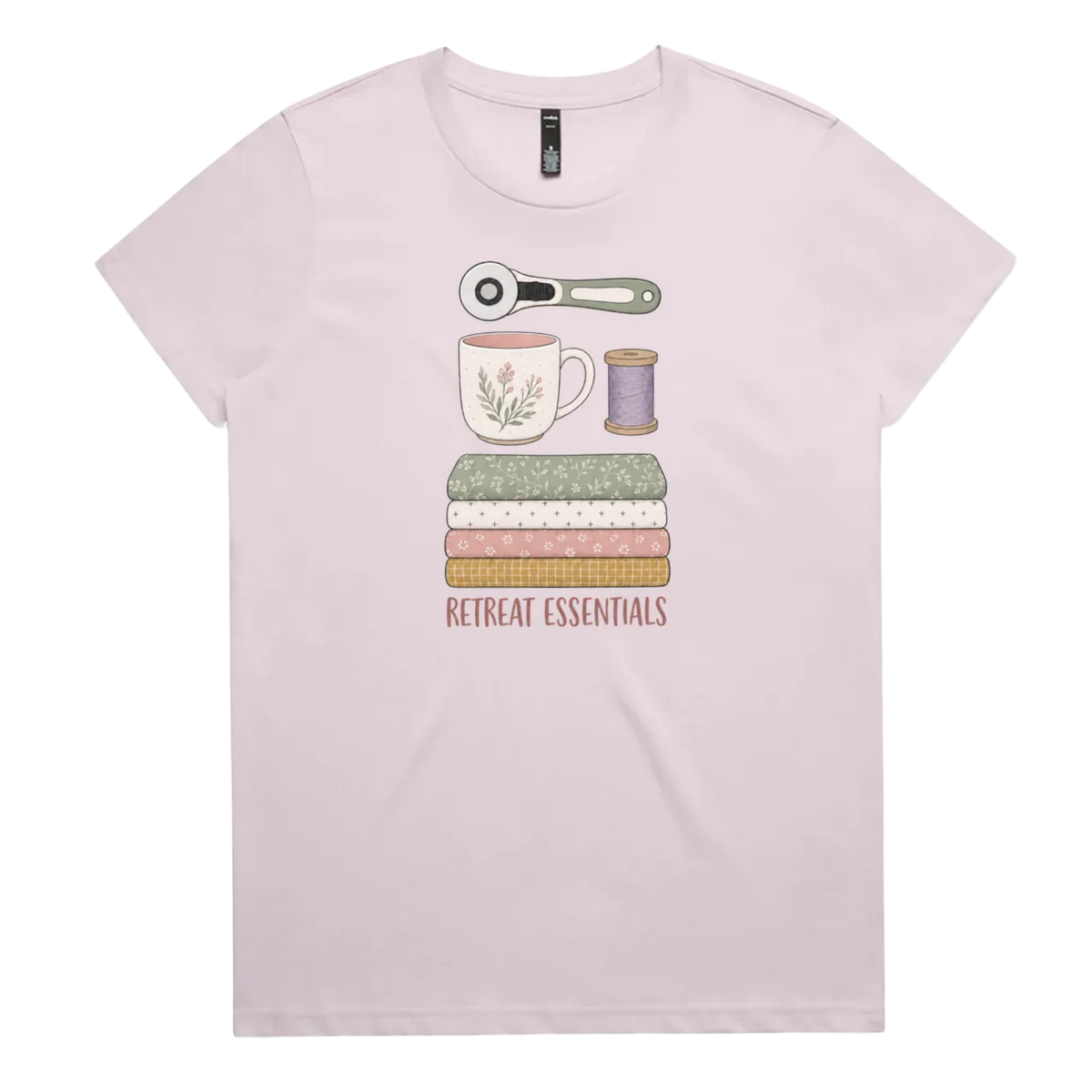 A light mauve short-sleeve crewneck t-shirt featuring a graphic of sewing and crafting essentials (a rotary cutter, mug, fabric stack, spool of thread) with the text 'RETREAT ESSENTIALS' printed below.