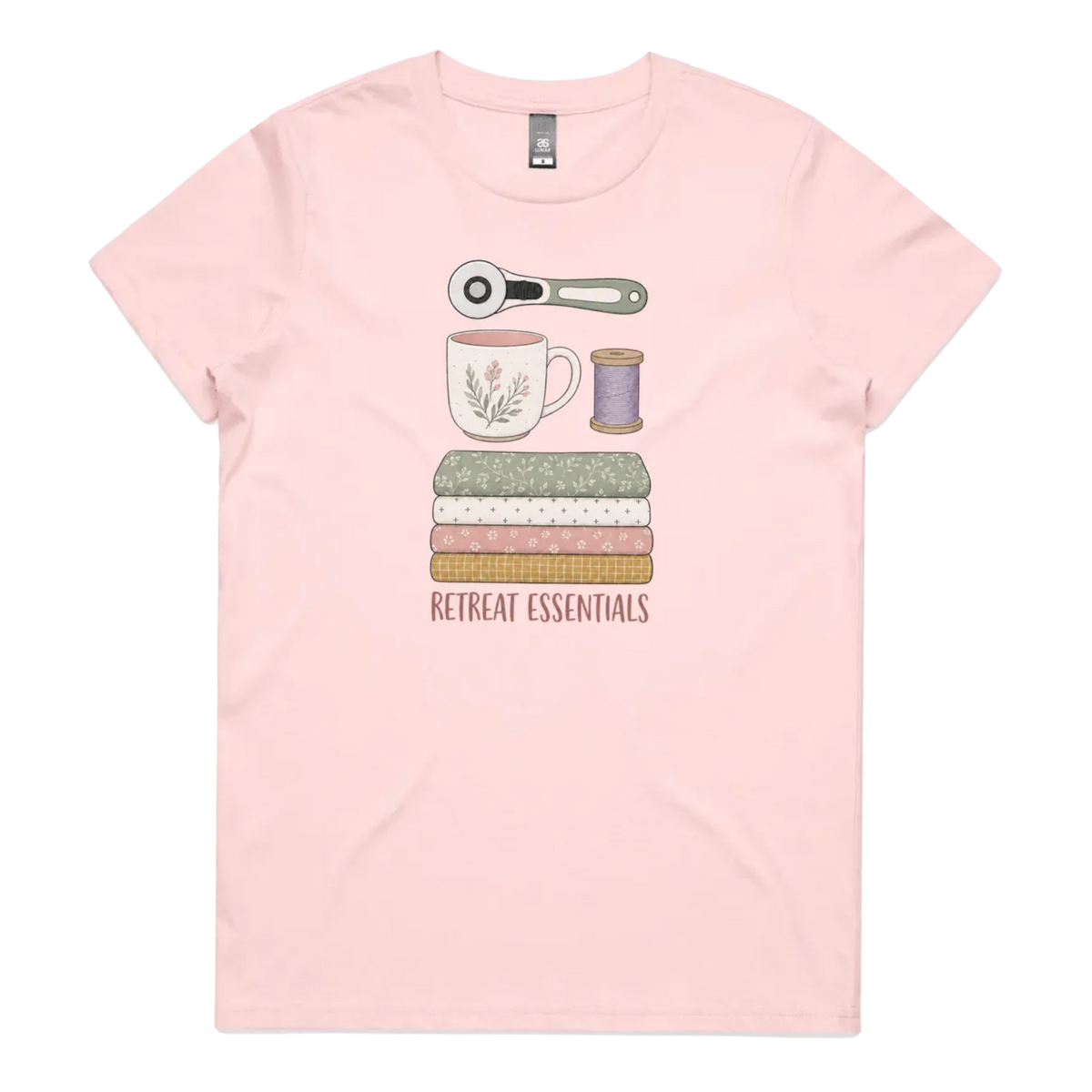 A light pink short-sleeve crewneck t-shirt featuring a graphic of sewing and crafting essentials (a rotary cutter, mug, fabric stack, spool of thread) with the text 'RETREAT ESSENTIALS' printed below.
