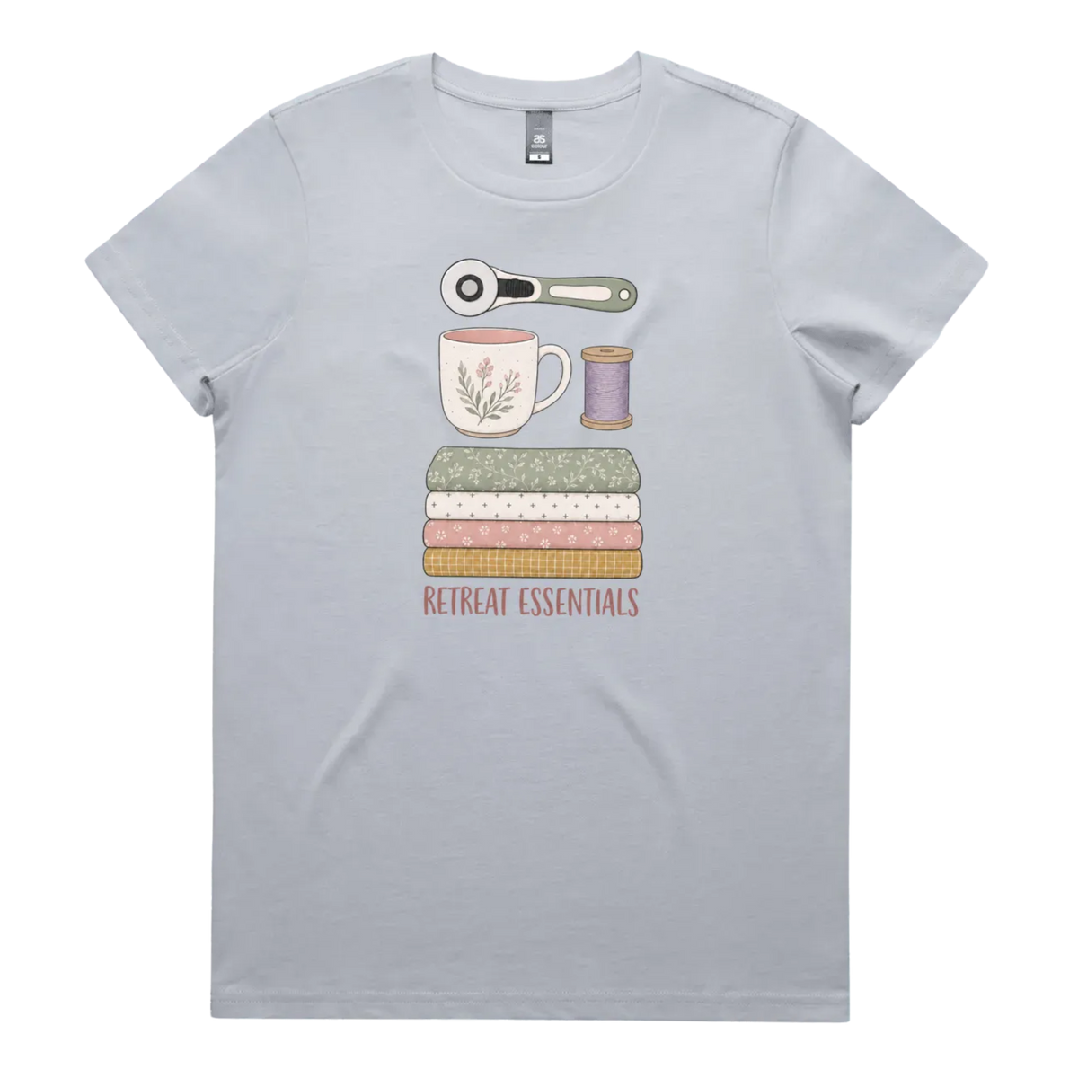 A powder blue short-sleeve crewneck t-shirt featuring a graphic of sewing and crafting essentials (a rotary cutter, mug, fabric stack, spool of thread) with the text 'RETREAT ESSENTIALS' printed below.