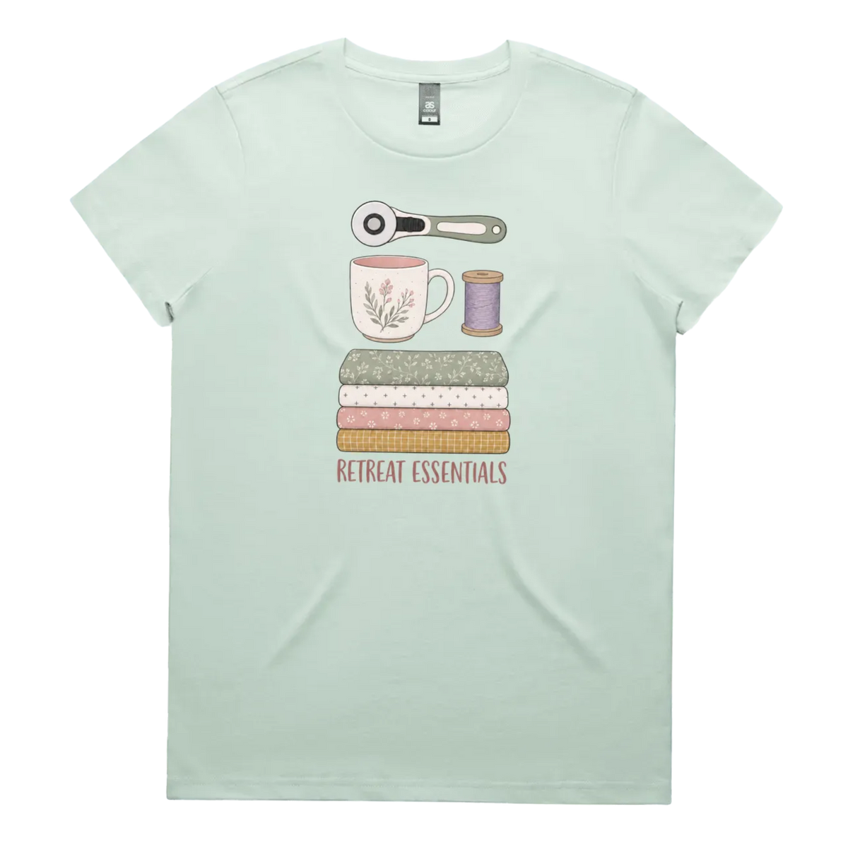 A light mint green short-sleeve crewneck t-shirt featuring a graphic of sewing and crafting essentials (a rotary cutter, mug, fabric stack, spool of thread) with the text 'RETREAT ESSENTIALS' printed below.