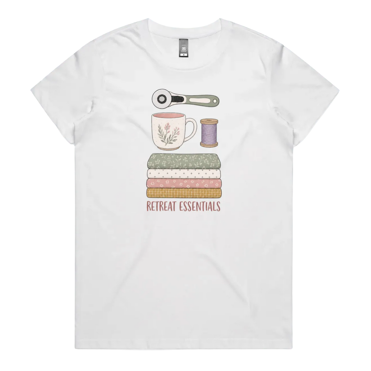 A white short-sleeve crewneck t-shirt featuring a graphic of sewing and crafting essentials (a rotary cutter, mug, fabric stack, spool of thread) with the text 'RETREAT ESSENTIALS' printed below.