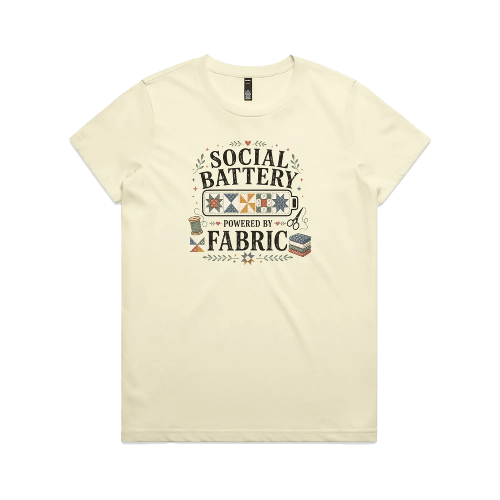 A pale butter yellow short-sleeve t-shirt with the graphic 'Social Battery Powered By Fabric'. Illustrations of patchwork blocks inside a battery, quilting tools and small leaves add decoration.