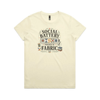 A pale butter yellow short-sleeve t-shirt with the graphic 'Social Battery Powered By Fabric'. Illustrations of patchwork blocks inside a battery, quilting tools and small leaves add decoration.
