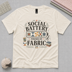 An off-white short-sleeve t-shirt laying flat on a pale grey backbround with quilting tools in the corners. The graphic on the t-shirt reads 'Social Battery Powered by Fabric' with an illustration of a battery filled with patchwork blocks, thread spool, scissors and fabric stack.
