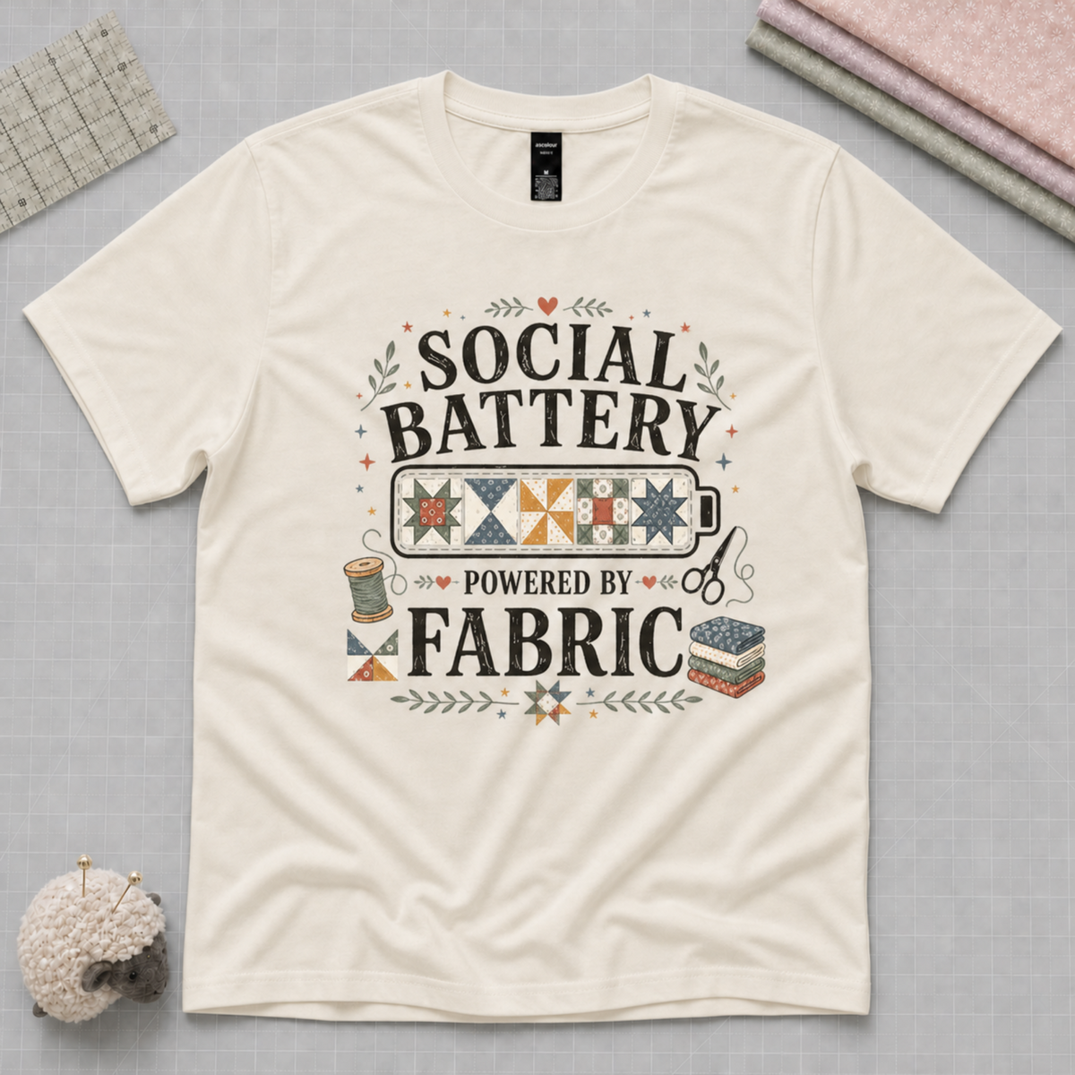An off-white short-sleeve t-shirt laying flat on a pale grey backbround with quilting tools in the corners. The graphic on the t-shirt reads 'Social Battery Powered by Fabric' with an illustration of a battery filled with patchwork blocks, thread spool, scissors and fabric stack.