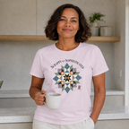 A woman with dark hair standing in a kitchen holding a cup of coffee. She is wearing a pale mauve graphic t-shirt with the words 'Scrappy but Sophisticated' above a large star quilt block.