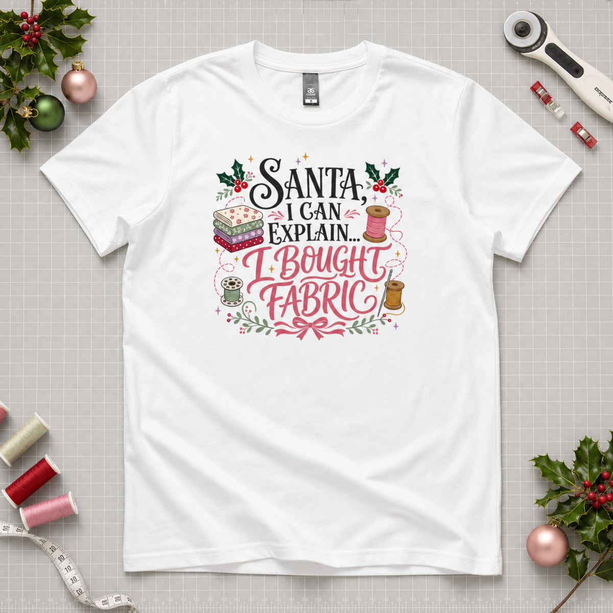 A white short-sleeve graphic T-shirt laid flat on a gray cutting mat, surrounded by  spools of thread, a rotary cutter, and holly. The shirt features a decorative printed slogan reading 'Santa, I Can Explain...I Bought Fabric'.