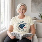 A smiling woman with short blonde hair sits in a light gray armchair, holding an open quilt pattern book. She is wearing a cream-colored graphic t-shirt that reads 'SLOW STITCHING CLUB' with a blue sashiko-stitched fabric illustration and the text 'Needle Thread - Time'.