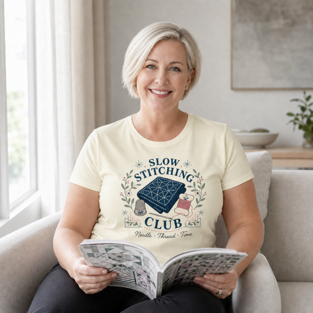 A smiling woman with short blonde hair sits in a light gray armchair, holding an open quilt pattern book. She is wearing a cream-colored graphic t-shirt that reads 'SLOW STITCHING CLUB' with a blue sashiko-stitched fabric illustration and the text 'Needle Thread - Time'.