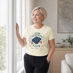 A woman with short blonde hair stands by a window in a bright room, wearing a pale yellow graphic t-shirt that reads 'SLOW STITCHING CLUB' with a sashiko-stitched fabric illustration, paired with black pants.
