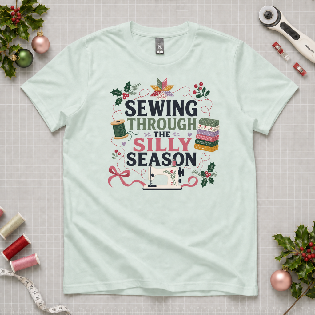 A mint green short-sleeve t-shirt with a festive graphic reading 'SEWING THROUGH THE SILLY SEASON' in black, green and pink lettering, surrounded by sewing and holiday motifs like a spool of thread, fabric stack, ribbon, holly leaves, and a sewing machine. The shirt is laid flat on a gridded cutting mat surrounded by craft props like spools of thread, a rotary cutter, and Christmas ornaments.