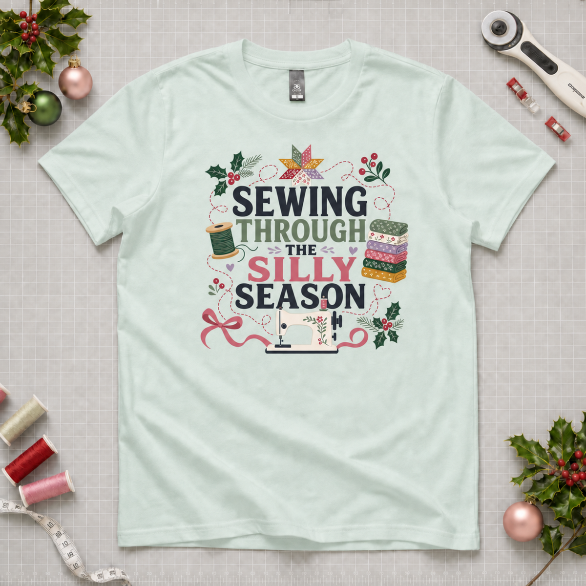 A mint green short-sleeve t-shirt with a festive graphic reading 'SEWING THROUGH THE SILLY SEASON' in black, green and pink lettering, surrounded by sewing and holiday motifs like a spool of thread, fabric stack, ribbon, holly leaves, and a sewing machine. The shirt is laid flat on a gridded cutting mat surrounded by craft props like spools of thread, a rotary cutter, and Christmas ornaments.
