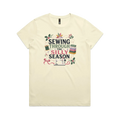 A pale butter yellow crewneck, short-sleeve t-shirt with the text'Sewing Through The Silly Season', surrounded by a sewing machine, fabric stack, ribbon, thread spool, a patchwork star and holly.