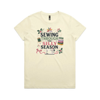 A pale butter yellow crewneck, short-sleeve t-shirt with the text'Sewing Through The Silly Season', surrounded by a sewing machine, fabric stack, ribbon, thread spool, a patchwork star and holly.