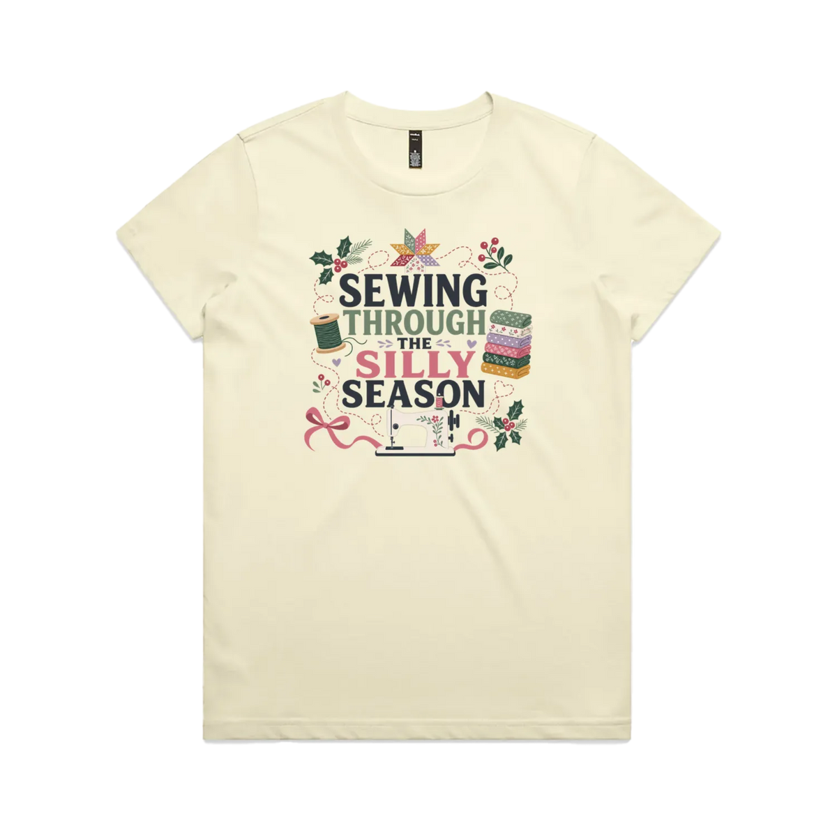 A pale butter yellow crewneck, short-sleeve t-shirt with the text'Sewing Through The Silly Season', surrounded by a sewing machine, fabric stack, ribbon, thread spool, a patchwork star and holly.