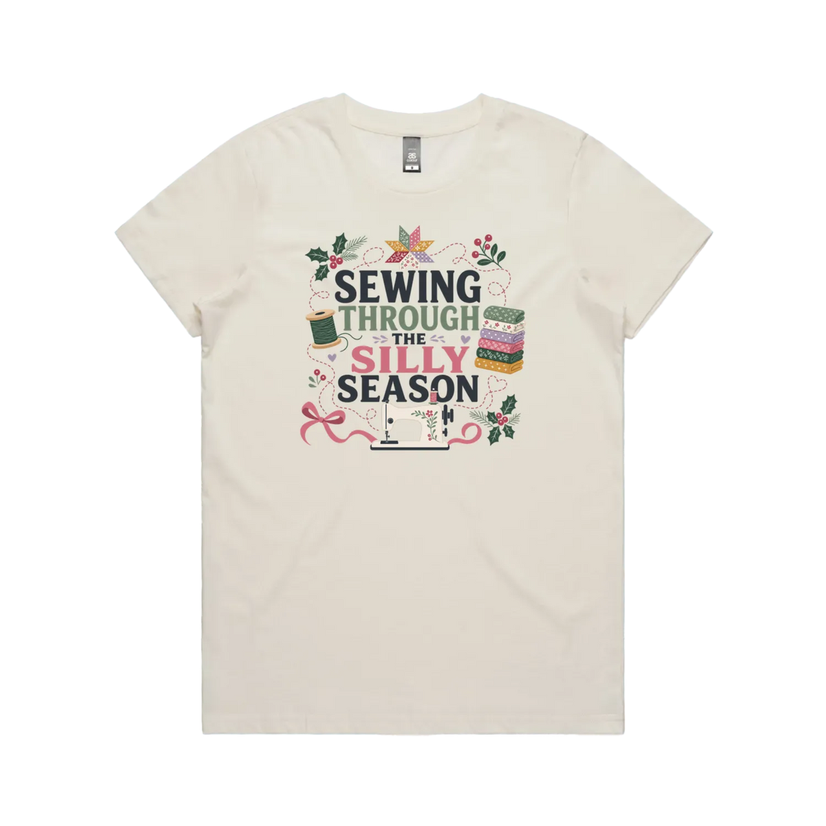 A pale cream crewneck, short-sleeve t-shirt with the text'Sewing Through The Silly Season', surrounded by a sewing machine, fabric stack, ribbon, thread spool, a patchwork star and holly.