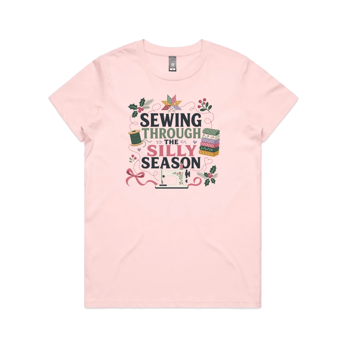 A pale pink crewneck, short-sleeve t-shirt with the text'Sewing Through The Silly Season', surrounded by a sewing machine, fabric stack, ribbon, thread spool, a patchwork star and holly.
