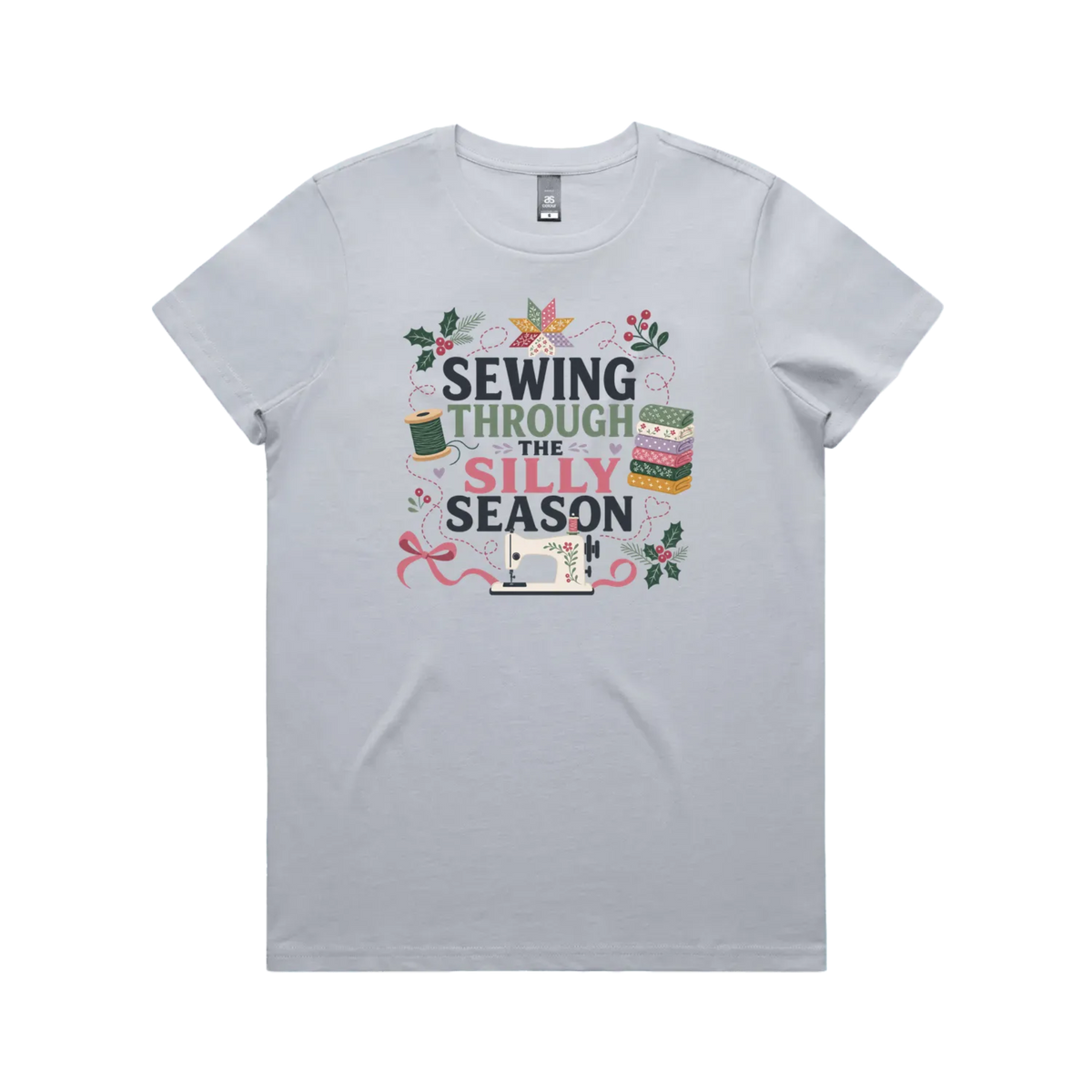 A powder blue crewneck, short-sleeve t-shirt with the text'Sewing Through The Silly Season', surrounded by a sewing machine, fabric stack, ribbon, thread spool, a patchwork star and holly.