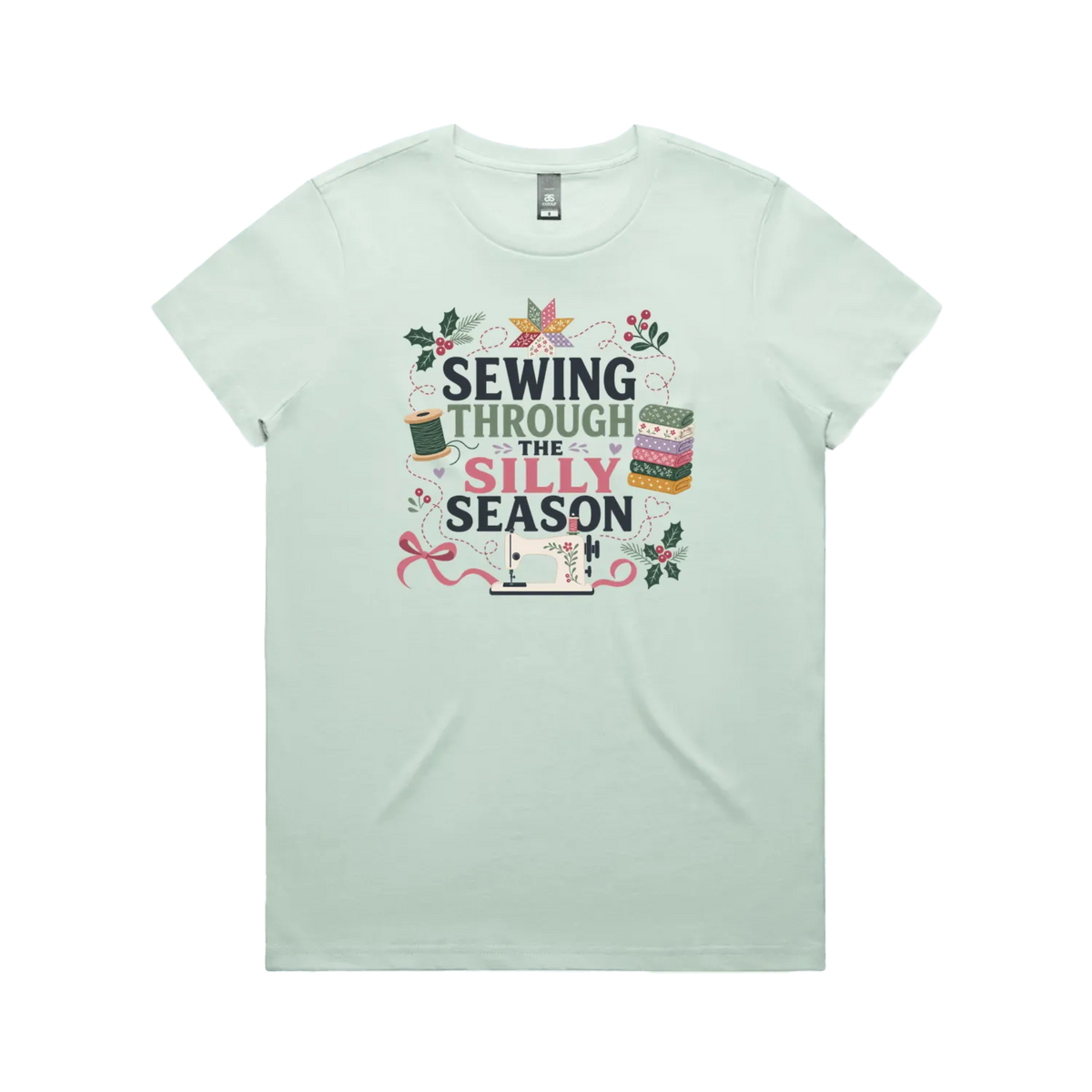 A pale seafoam green crewneck, short-sleeve t-shirt with the text'Sewing Through The Silly Season', surrounded by a sewing machine, fabric stack, ribbon, thread spool, a patchwork star and holly.