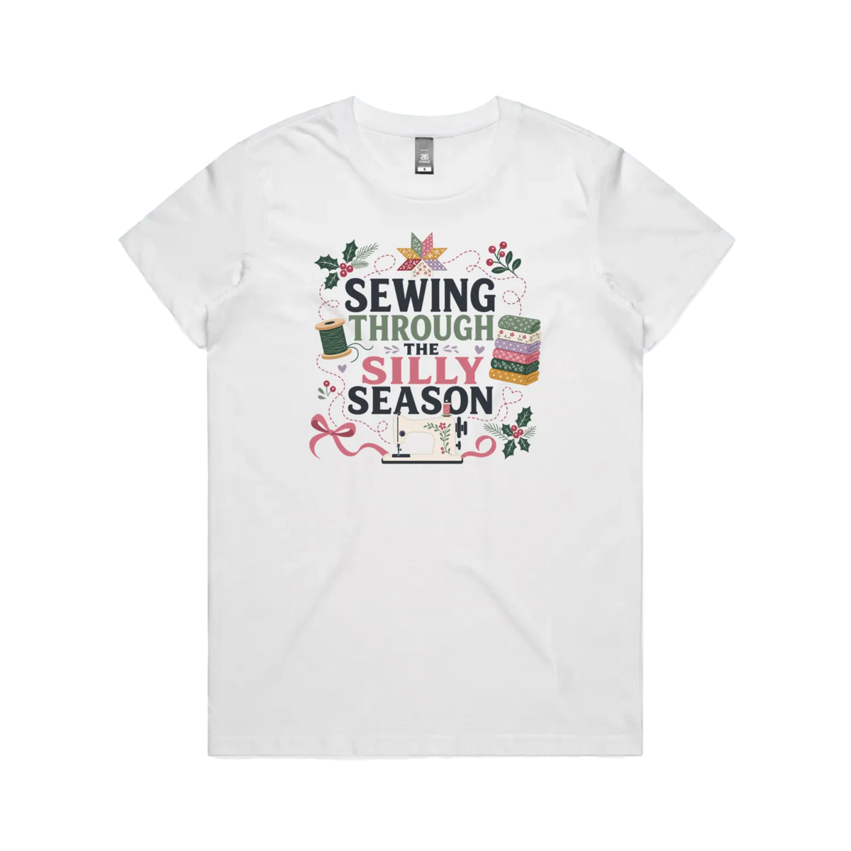 A white crewneck, short-sleeve t-shirt with the text'Sewing Through The Silly Season', surrounded by a sewing machine, fabric stack, ribbon, thread spool, a patchwork star and holly.