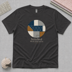 A dark charcoal gray t-shirt laid flat on a textured gray background, featuring a circular graphic of stitched fabric patterns in blue, beige, and brown, with the text 'Stitched With Intention' printed below. Folded fabric, quilt ruler and pincushion are visible in the corners.