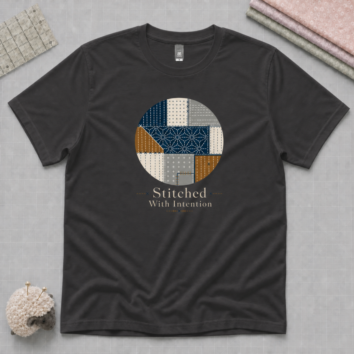 A dark charcoal gray t-shirt laid flat on a textured gray background, featuring a circular graphic of stitched fabric patterns in blue, beige, and brown, with the text 'Stitched With Intention' printed below. Folded fabric, quilt ruler and pincushion are visible in the corners.