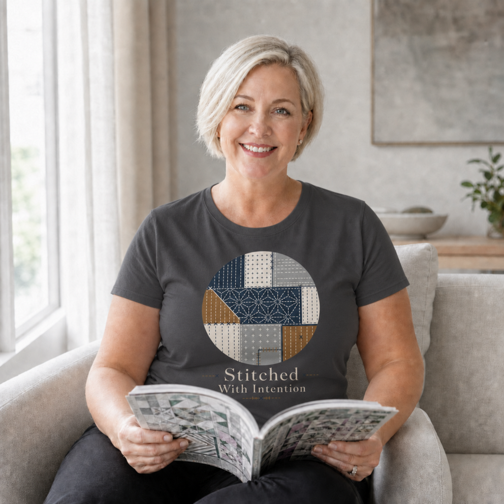 A smiling woman with short blonde hair sits in a beige armchair, holding an open book. She is wearing a dark charcoal gray T-shirt with a circular quilt-pattern graphic and the text 'Stitched With Intention' on the front.