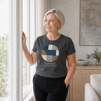 A woman with short blonde hair stands by a window in a bright room, wearing a dark charcoal gray t-shirt with a circular patchwork graphic in blue, beige, and white, and the text 'Stitched With Intention' printed below it.