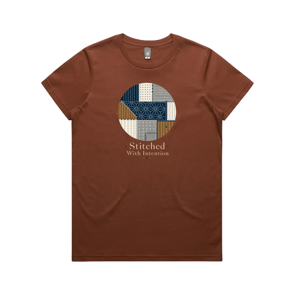 A clay brown short-sleeve crewneck t-shirt featuring a circular graphic on the chest with geometric sashiko stitched patterns in navy, beige, and brown, with the text 'Stitched With Intention' printed below.