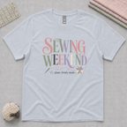A powder blue short-sleeve t-shirt laying flat on a pale grey background with quilting tools in the corners. The graphic reads 'Sewing Weekend plans firmly made' in muted pink, green, mauve, grey and cream.