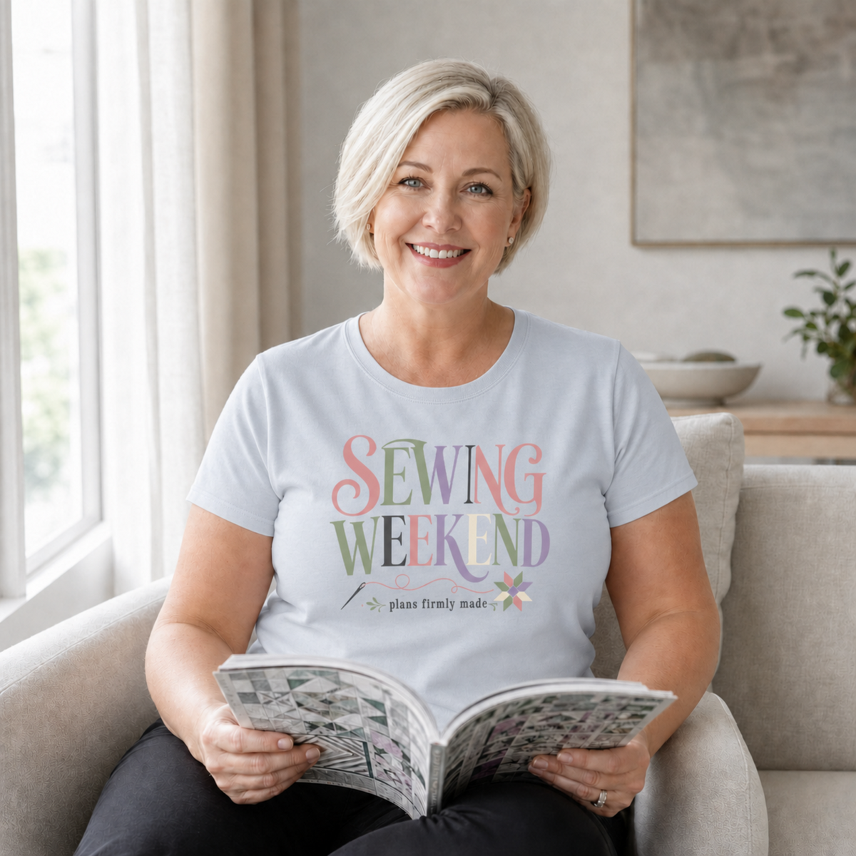 A smiling blond woman sits on a beige lounge holding an open book. She is wearing a powder blue short-sleeve t-shirt with the words 'Sewing Weekend plans firmly made' in pink, green, mauve, grey and cream.