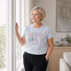 A smiling blond woman stands looking out a window in a lounge room. She is wearing dark jeans and a powder blue short-sleeve t-shirt with the words 'Sewing Weekend plans firmly made' in pink, green, mauve, grey and cream.