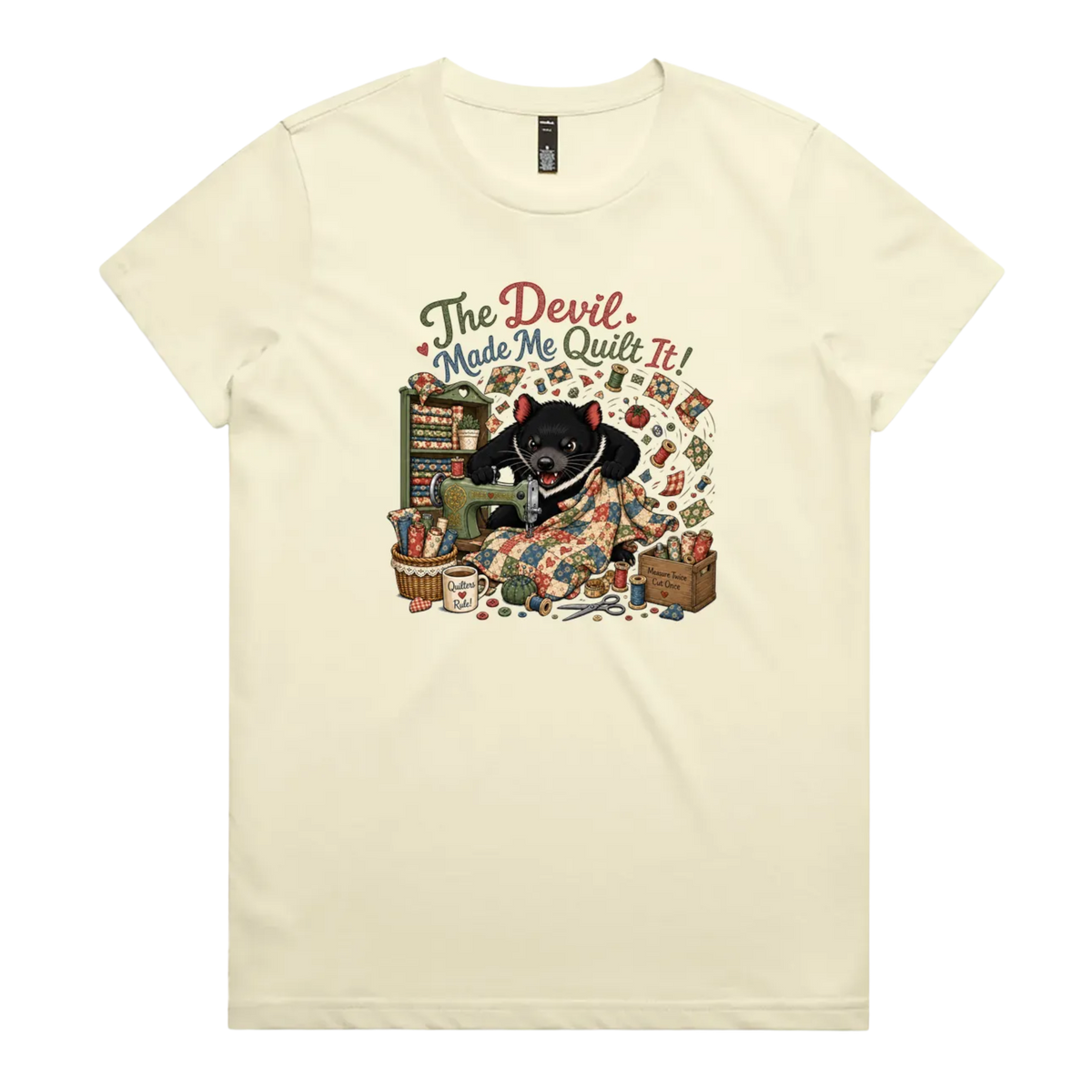 A light butter yellow short-sleeve crewneck t-shirt featuring a colorful graphic print of a Tasmanian Devil surrounded by fabric, a mug, a basket and wooden crate with fabric rolls, and the text 'The Devil Made Me Quilt It!' in a retro font.