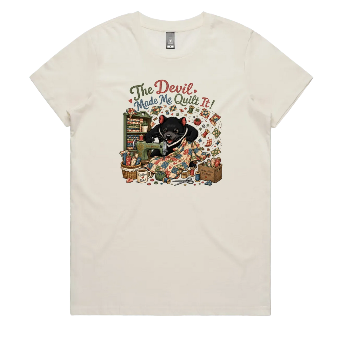 A light cream short-sleeve crewneck t-shirt featuring a colorful graphic print of a Tasmanian Devil surrounded by fabric, a mug, a basket and wooden crate with fabric rolls, and the text 'The Devil Made Me Quilt It!' in a retro font.