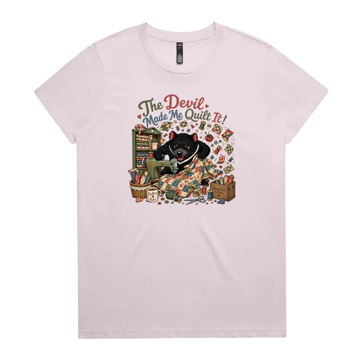 A light mauve short-sleeve crewneck t-shirt featuring a colorful graphic print of a Tasmanian Devil surrounded by fabric, a mug, a basket and wooden crate with fabric rolls, and the text 'The Devil Made Me Quilt It!' in a retro font.