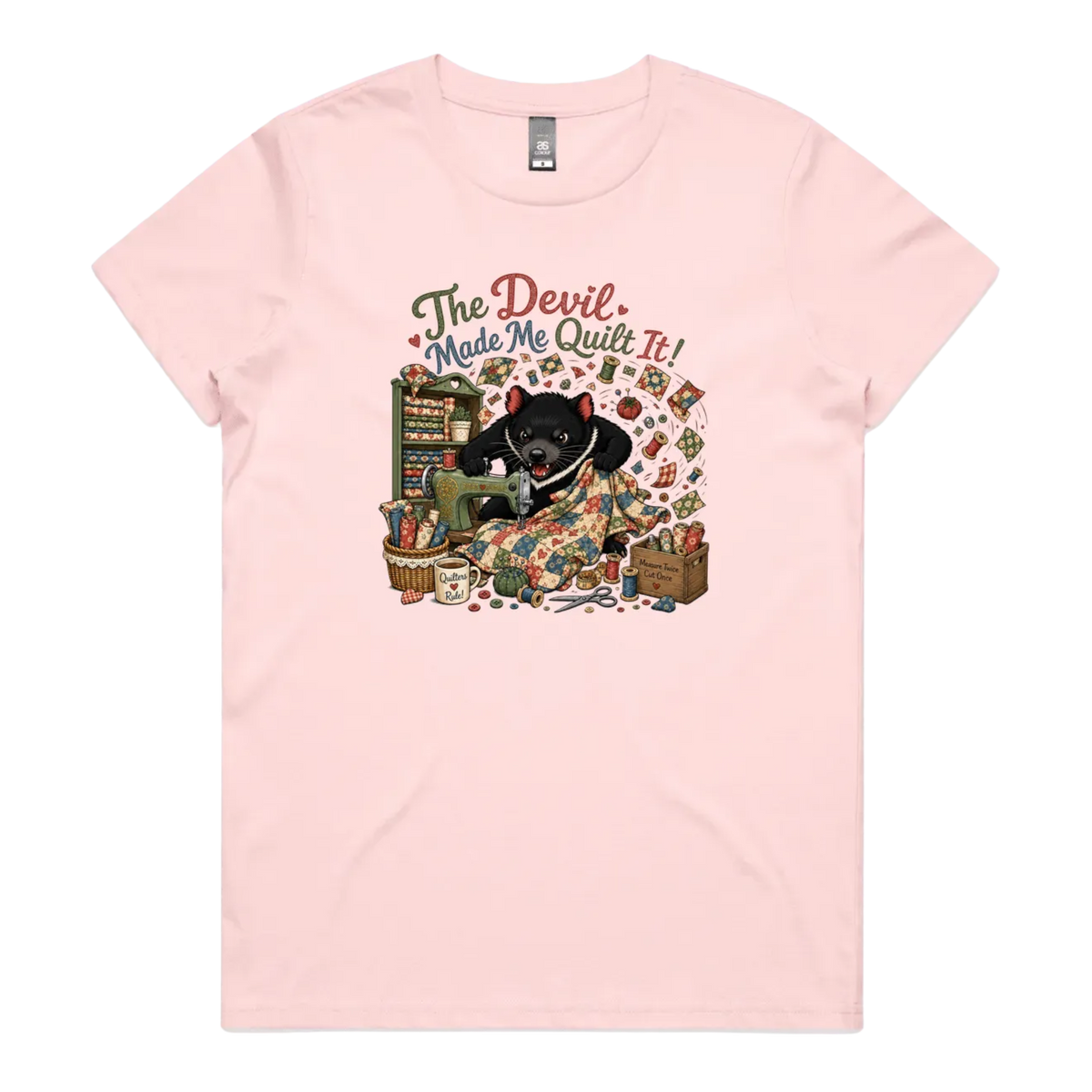 A light pink short-sleeve crewneck t-shirt featuring a colorful graphic print of a Tasmanian Devil surrounded by fabric, a mug, a basket and wooden crate with fabric rolls, and the text 'The Devil Made Me Quilt It!' in a retro font.