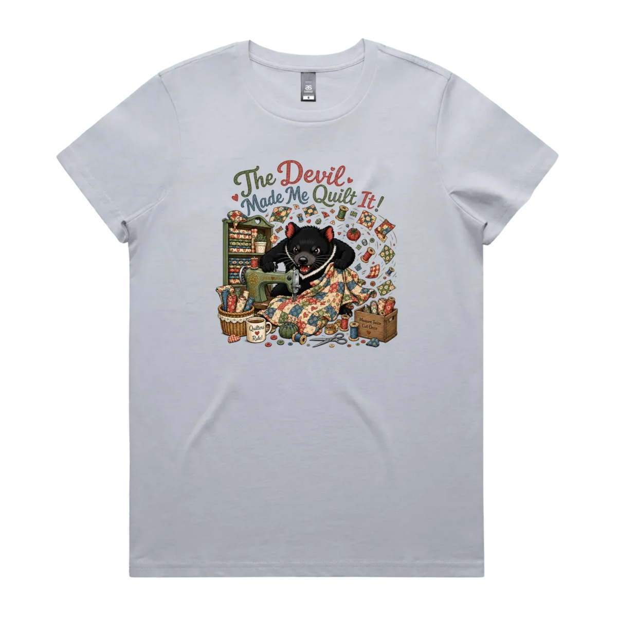 A light powder blue short-sleeve crewneck t-shirt featuring a colorful graphic print of a Tasmanian Devil surrounded by fabric, a mug, a basket and wooden crate with fabric rolls, and the text 'The Devil Made Me Quilt It!' in a retro font.