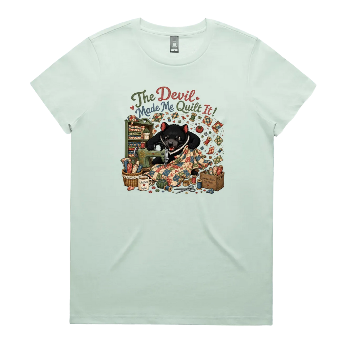 A light seafoam green short-sleeve crewneck t-shirt featuring a colorful graphic print of a Tasmanian Devil surrounded by fabric, a mug, a basket and wooden crate with fabric rolls, and the text 'The Devil Made Me Quilt It!' in a retro font.