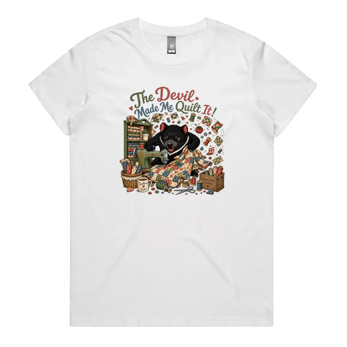 A white short-sleeve crewneck t-shirt featuring a colorful graphic print of a Tasmanian Devil surrounded by fabric, a mug, a basket and wooden crate with fabric rolls, and the text 'The Devil Made Me Quilt It!' in a retro font.