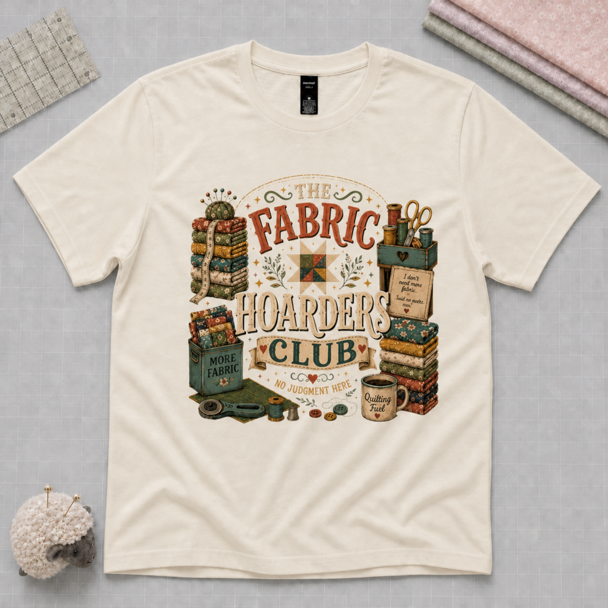 A pale cream short-sleeve graphic t-shirt featuring the words 'The Fabric Hoarders Club' surrounded by fabric bundles and quilting tools. The shirt lays flat on a pale grey cutting mage with fabric, a ruler and pincushion in the corners.