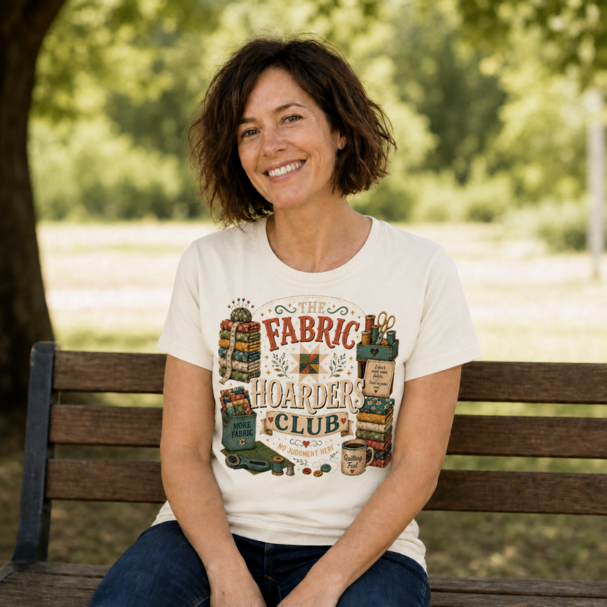 A smiling woman with dark hair sits on a bench in a part wearing a graphic t-shirt featuring the words 'The Fabric Hoarders Club' with illustrations of fabric bundles and quilting tools.