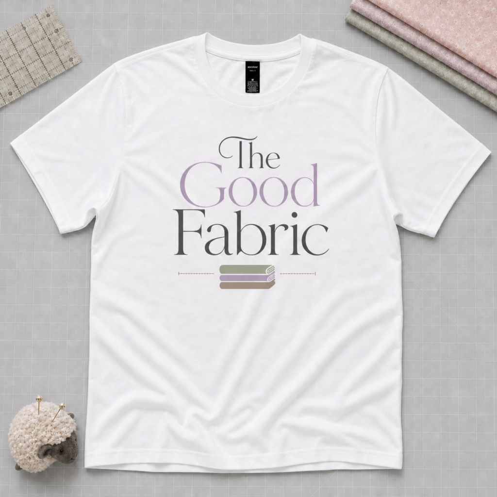 A white short-sleeve t-shirt laid flat on a pale grey cutting mat, featuring the text 'The Good Fabric' in black and lavender lettering with a small stack of fabric beneath. A quilting ruler, fabric stack and a sheep-shaped pincusion and visible in the corners.