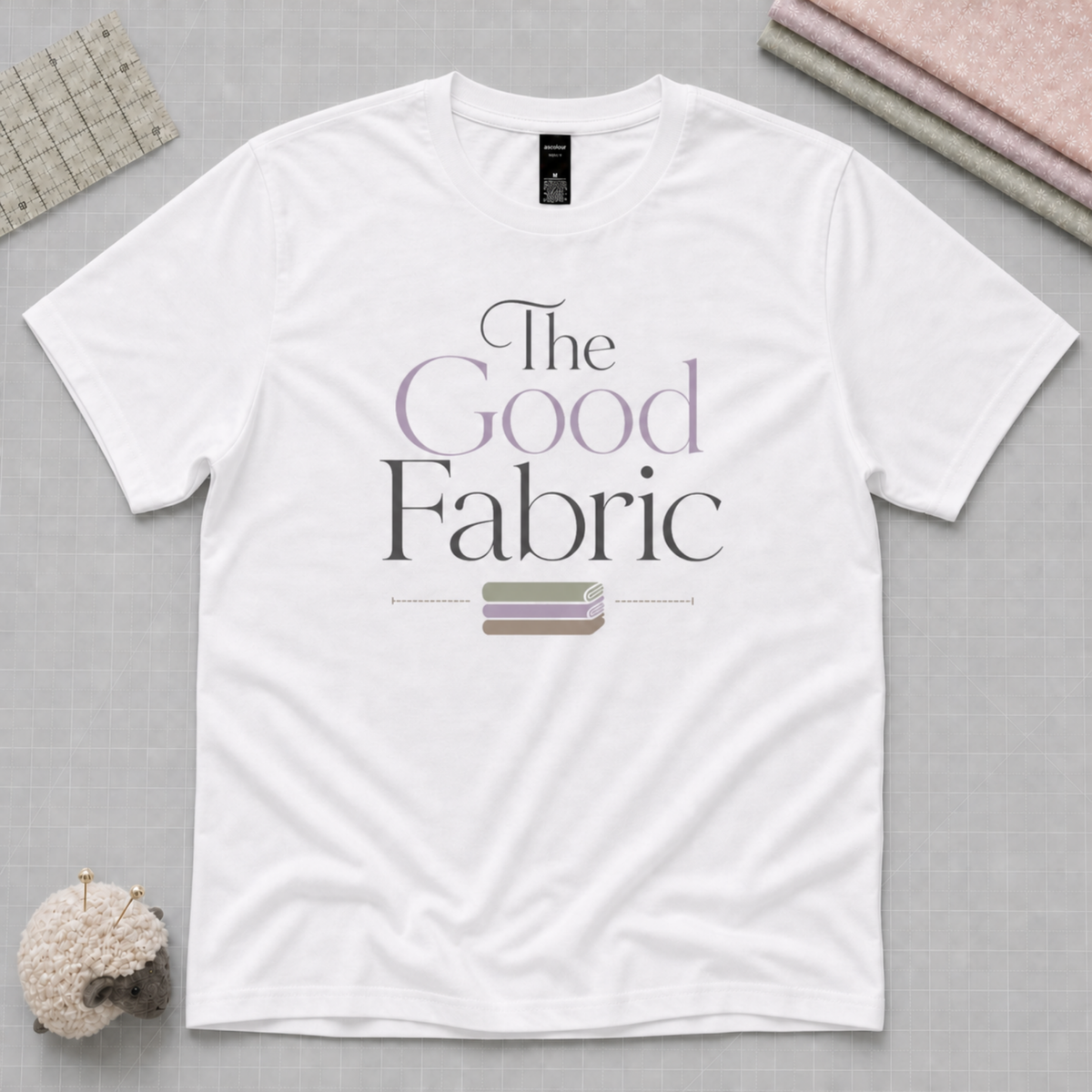 A white short-sleeve t-shirt laid flat on a pale grey cutting mat, featuring the text 'The Good Fabric' in black and lavender lettering with a small stack of fabric beneath. A quilting ruler, fabric stack and a sheep-shaped pincusion and visible in the corners.